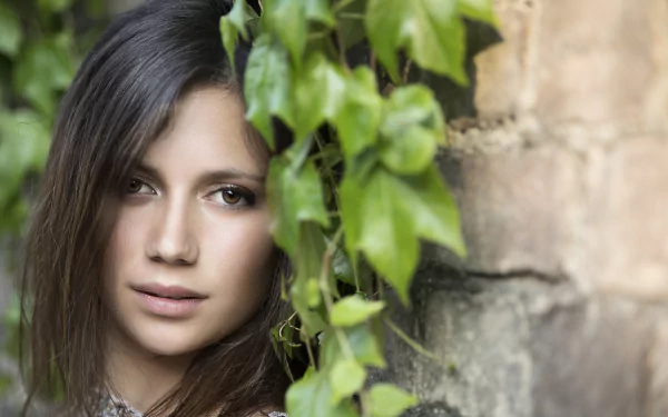 HD desktop wallpaper featuring a brunette model with brown eyes, posing next to an old brick wall adorned with green ivy.