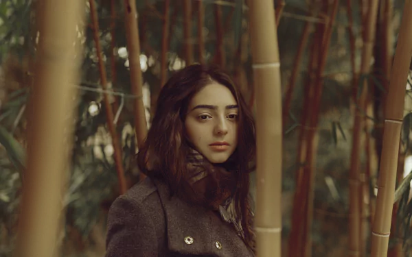 Brunette woman with brown eyes wrapped in a scarf and coat gazes pensively among bamboo stalks — moody outdoor HD desktop wallpaper.