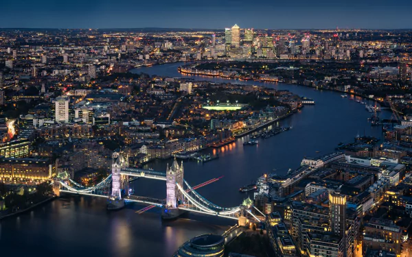 Night aerial view of London: illuminated Tower Bridge spanning the Thames, riverside buildings and distant horizon — 5K Ultra HD PC wallpaper of a man-made cityscape in the United Kingdom.