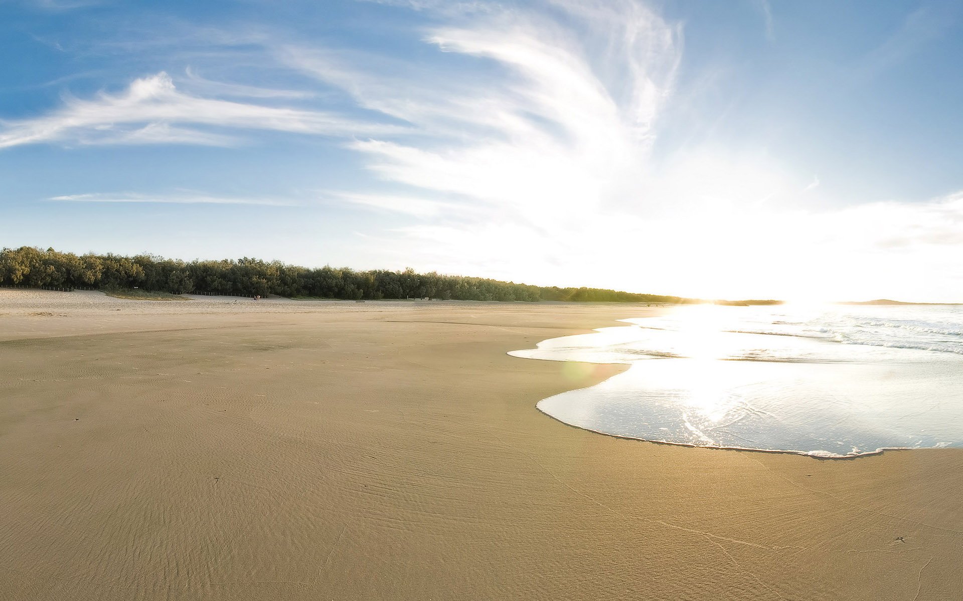 HD desktop wallpaper showcasing a serene beach with smooth sand, calm water reflecting sunlight, and a bright sky filled with wispy clouds, highlighting nature's beauty.