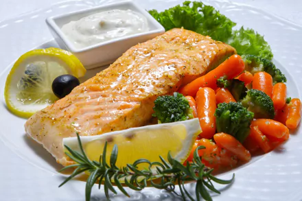 A vibrant meal featuring a cooked salmon fillet served with steamed carrots, broccoli, lemon wedges, a sprig of rosemary, lettuce, and a side of creamy sauce, captured in HD.