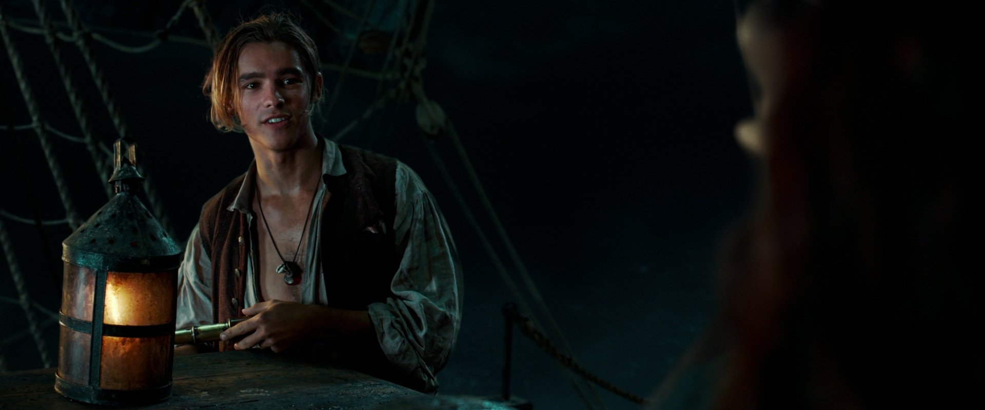 Dim 2K desktop wallpaper: a young sailor on a ship's deck at night, speaking to someone off-frame — scene from Pirates of the Caribbean: Dead Men Tell No Tales.
