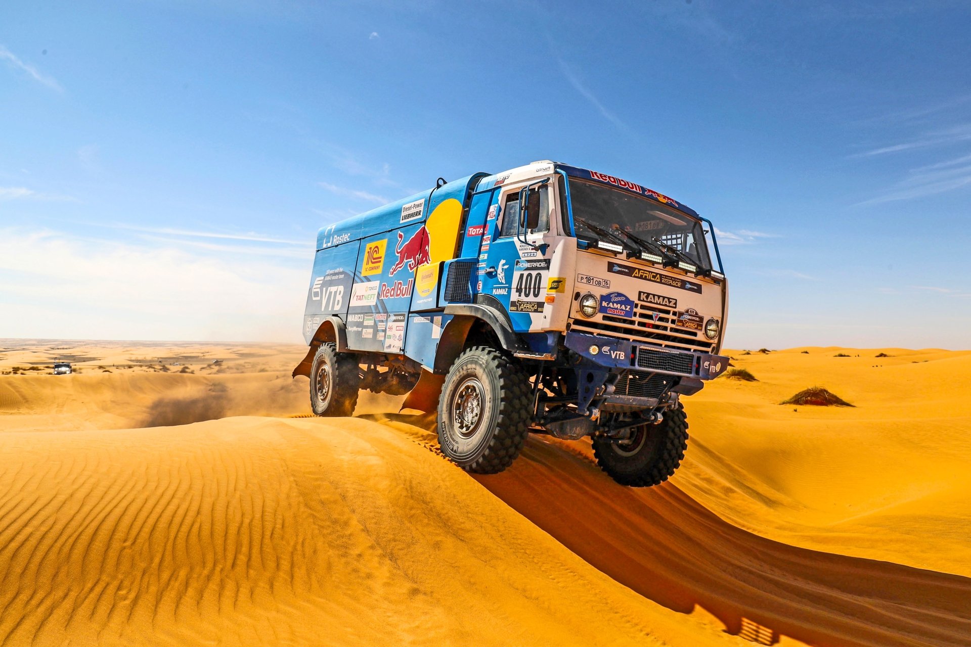 A Red Bull Kamaz rally truck racing across golden sand dunes under a clear blue horizon in a desert, captured in high-resolution 4K Ultra HD.