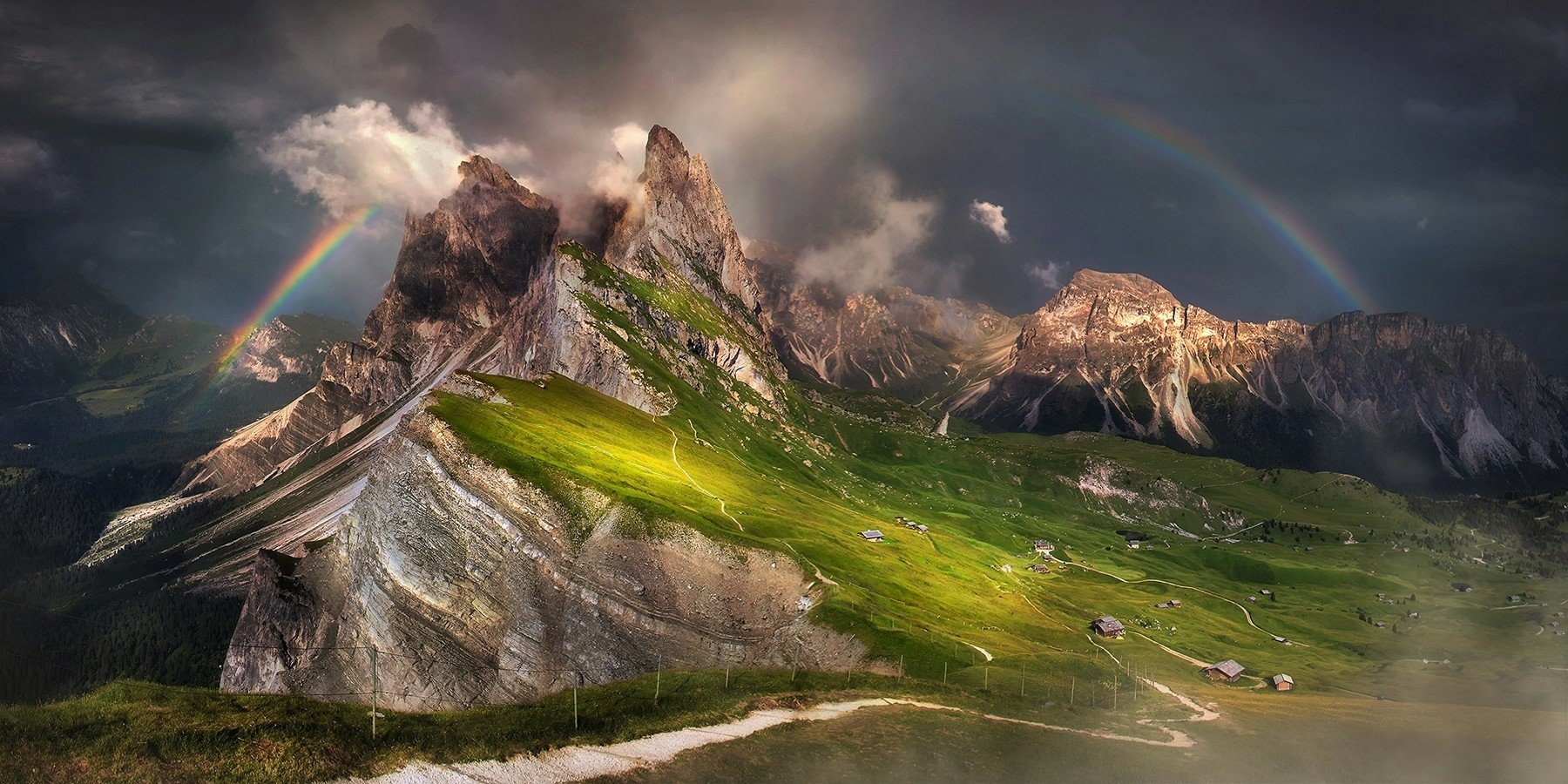 HD desktop wallpaper showing a mountain peak with scattered houses, surrounded by clouds, fog, and a vibrant rainbow arching across the natural landscape.