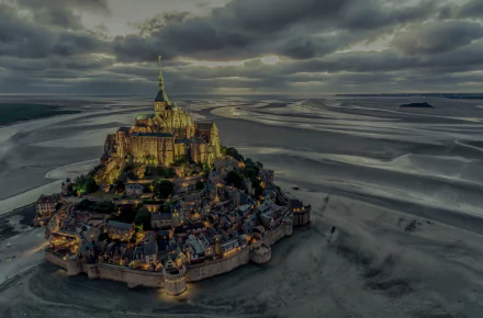 A dramatic HD desktop wallpaper of Mont Saint-Michel monastery on sandy horizon under cloudy skies in France, showcasing a stunning religious landmark.