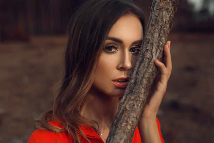 HD PC desktop wallpaper of a brunette woman model—close-up face with brown eyes, partially hidden by a tree branch, shallow depth of field and warm earthy backdrop.