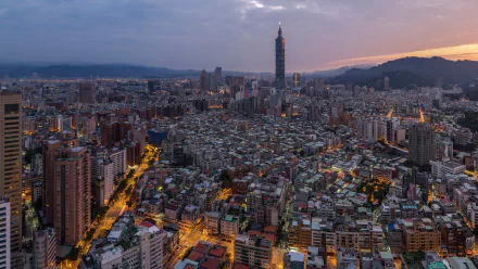 An HD wallpaper featuring the cityscape of Taipei at dusk, showcasing the iconic Taipei 101 skyscraper amidst a dense cluster of buildings, with mountains in the backdrop.