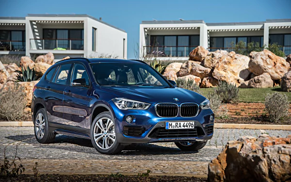 A blue BMW X1 SUV parked on a stone driveway with modern houses and large rocks in the background, captured in 4K Ultra HD for a PC desktop wallpaper.