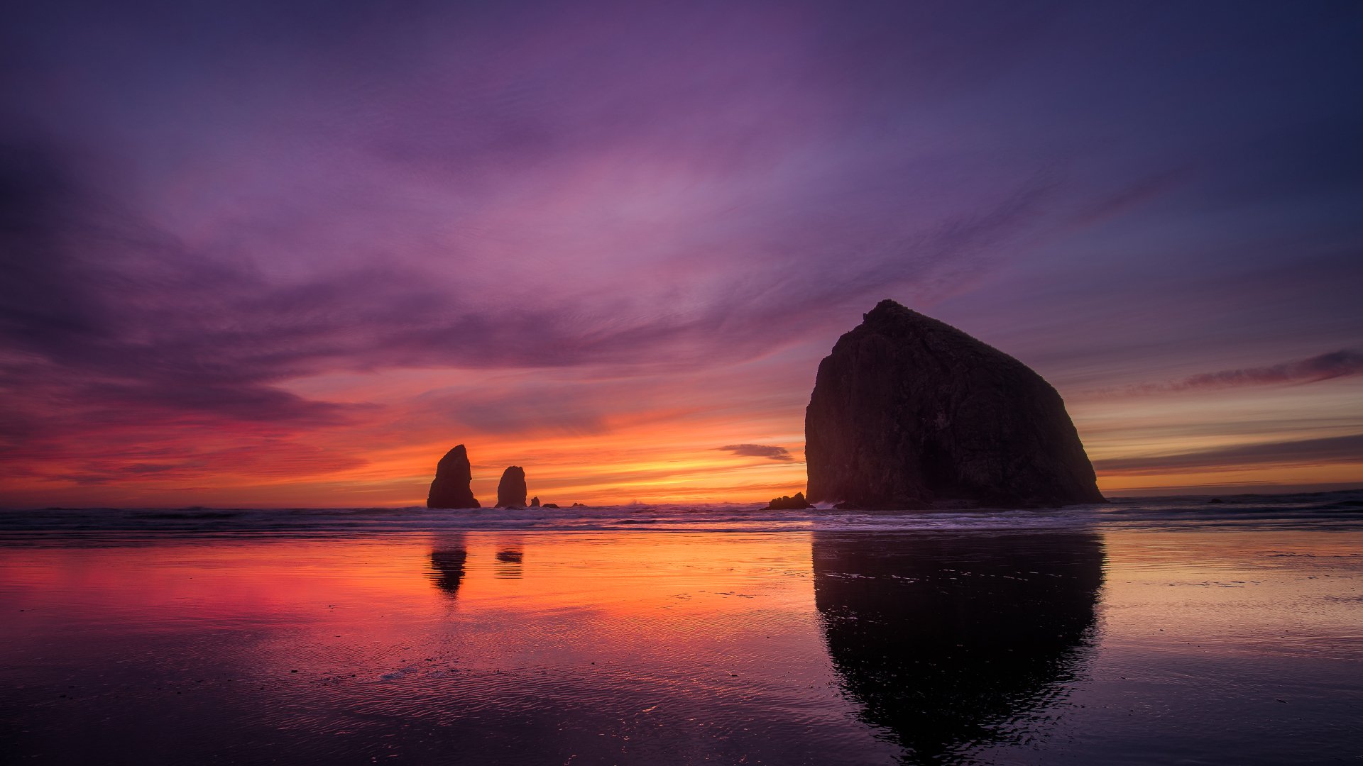 HD desktop wallpaper of a serene beach at sunset with large rocks silhouetted against a vibrant sky of purples, oranges, and pinks, reflecting beautifully over the calm water.