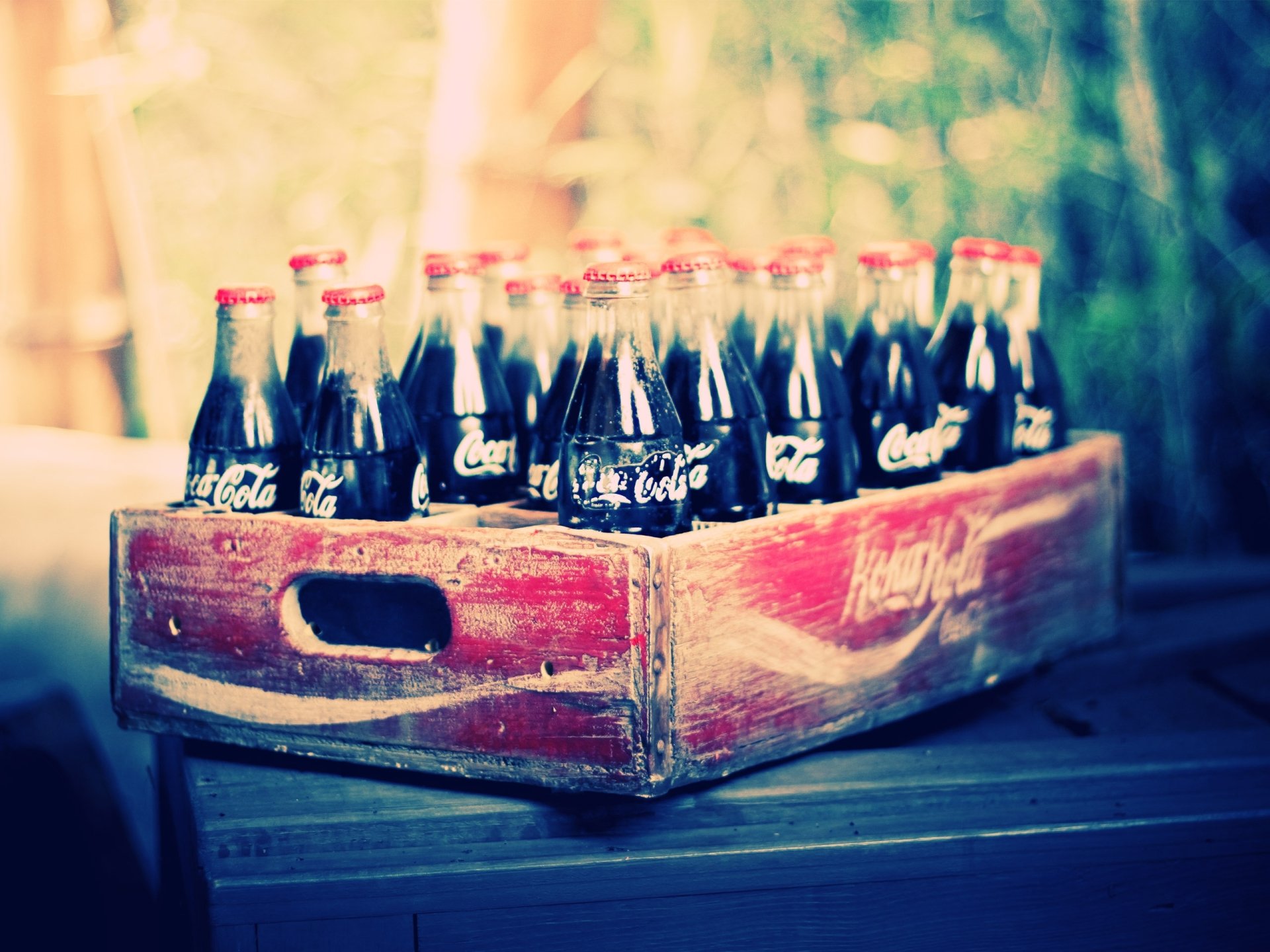 A high-definition desktop wallpaper featuring a vintage crate filled with Coca-Cola soda bottles, set against a blurred natural background.