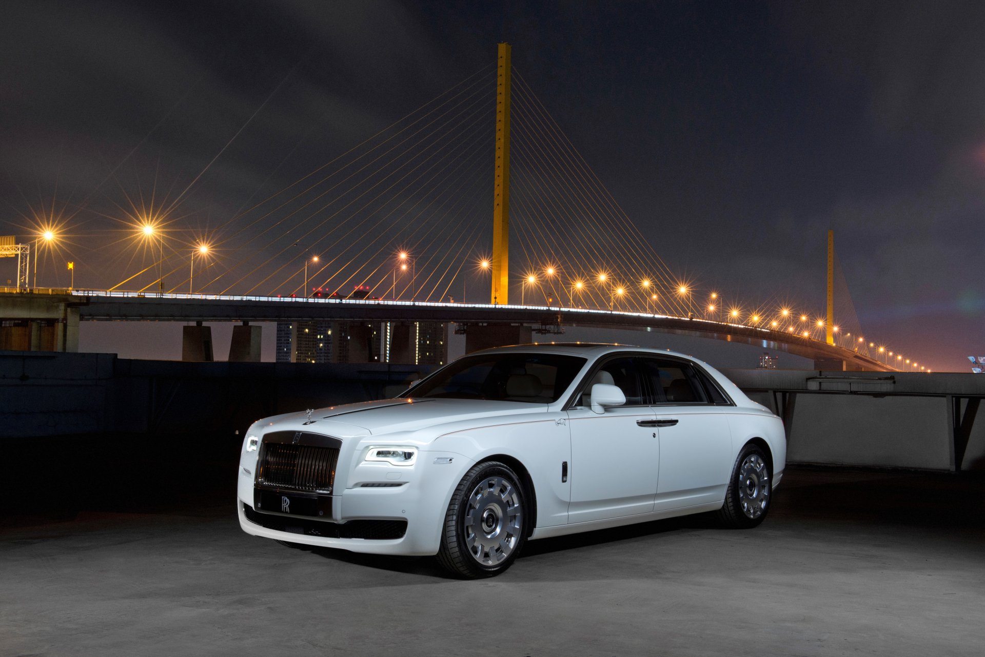 A white Rolls-Royce Ghost parked at night near a lit bridge, showcased in 4K Ultra HD as a sleek luxury vehicle against a dark sky.