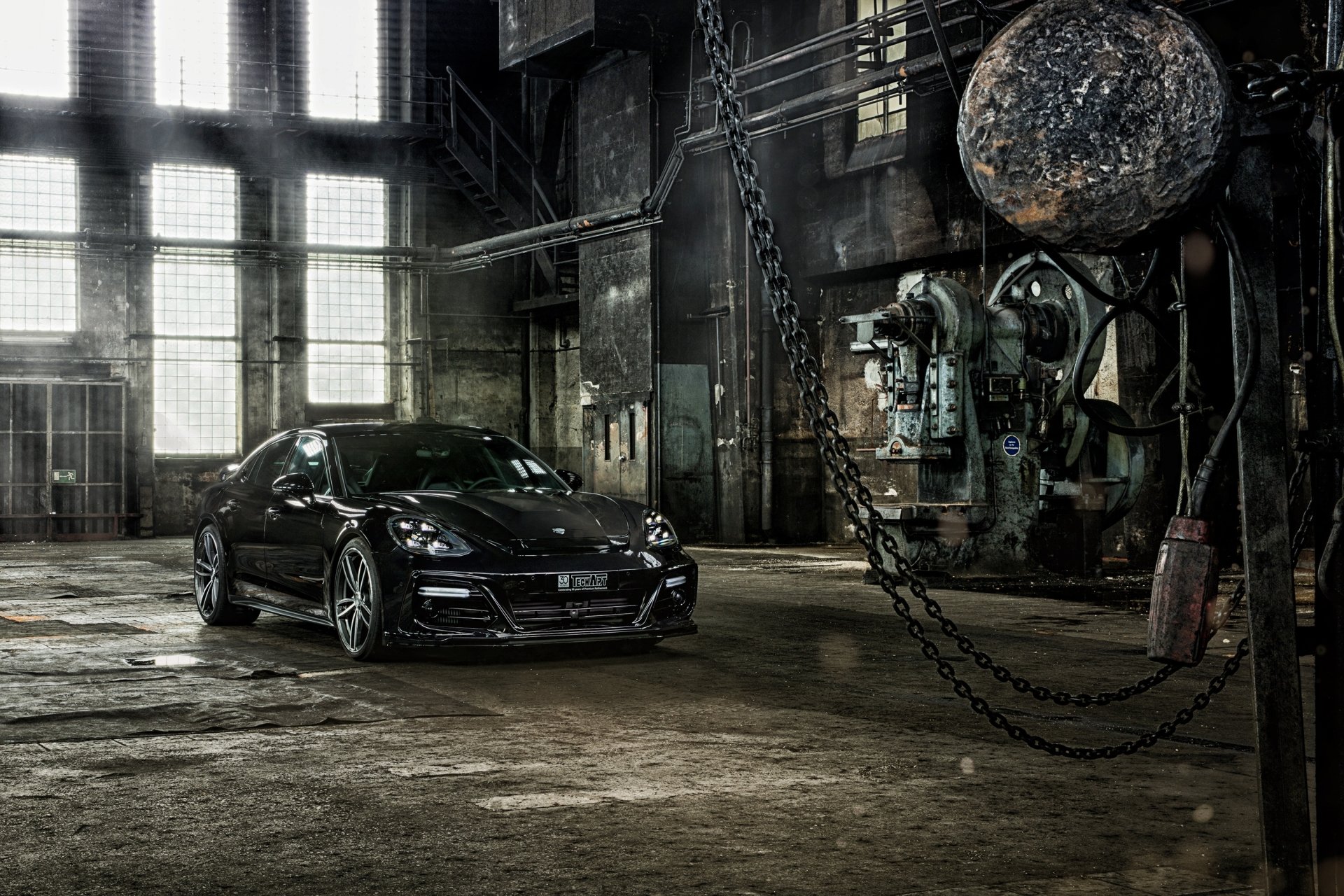 Black Porsche Panamera parked inside an abandoned factory with large windows, industrial machinery, and a rugged, gritty atmosphere in this HD PC desktop wallpaper.
