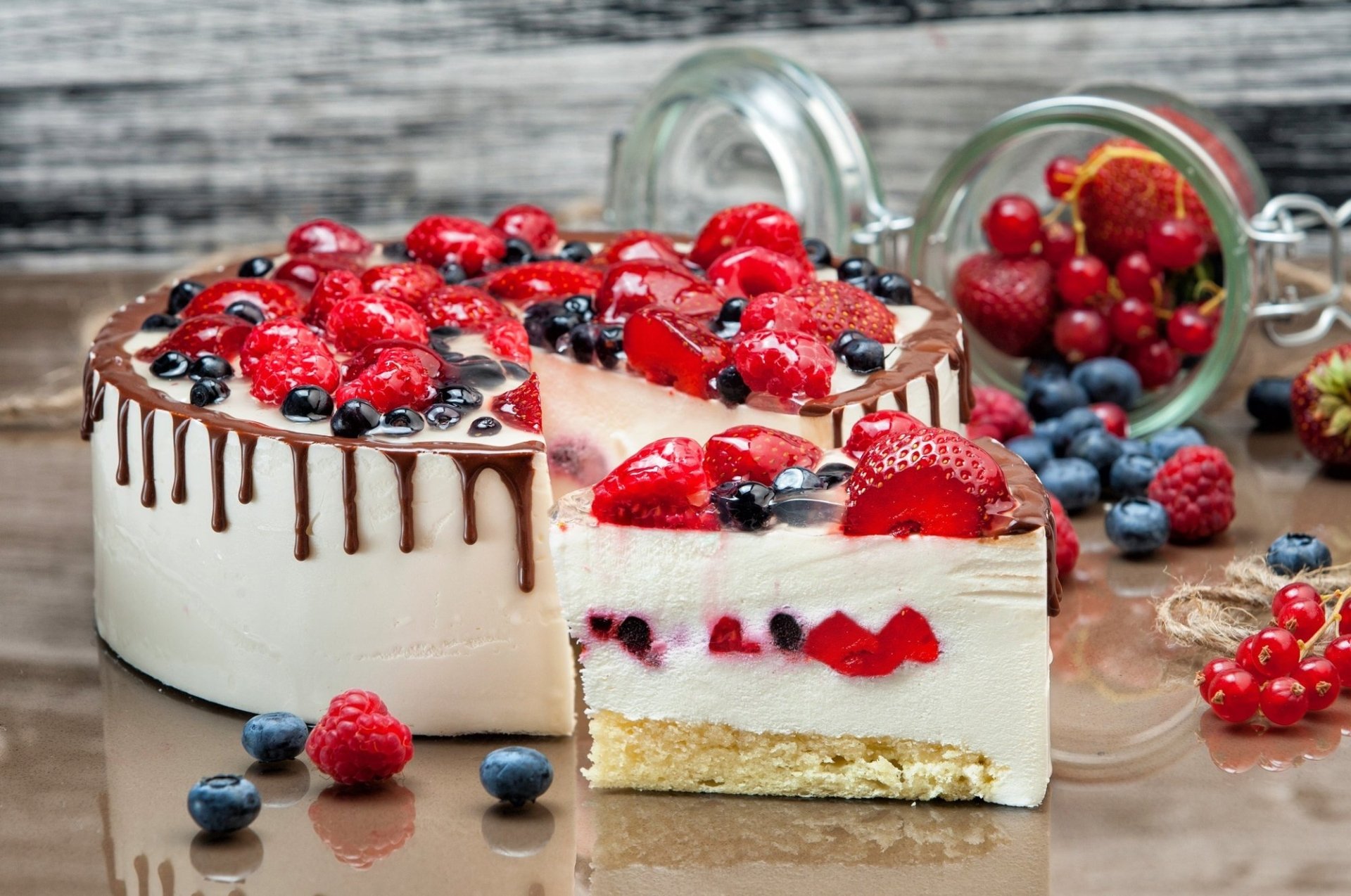 HD desktop wallpaper of a creamy cheesecake topped with strawberries, raspberries, blueberries, and chocolate drizzle, surrounded by fresh berries on a rustic wooden surface.