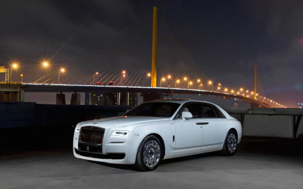 A white Rolls-Royce Ghost parked at night near a lit bridge, showcased in 4K Ultra HD as a sleek luxury vehicle against a dark sky.
