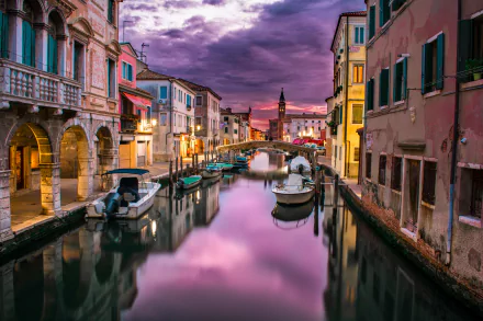 A stunning HD wallpaper of a sunset in Chioggia, Venice, featuring colorful houses, boats in a calm canal, and their reflections.