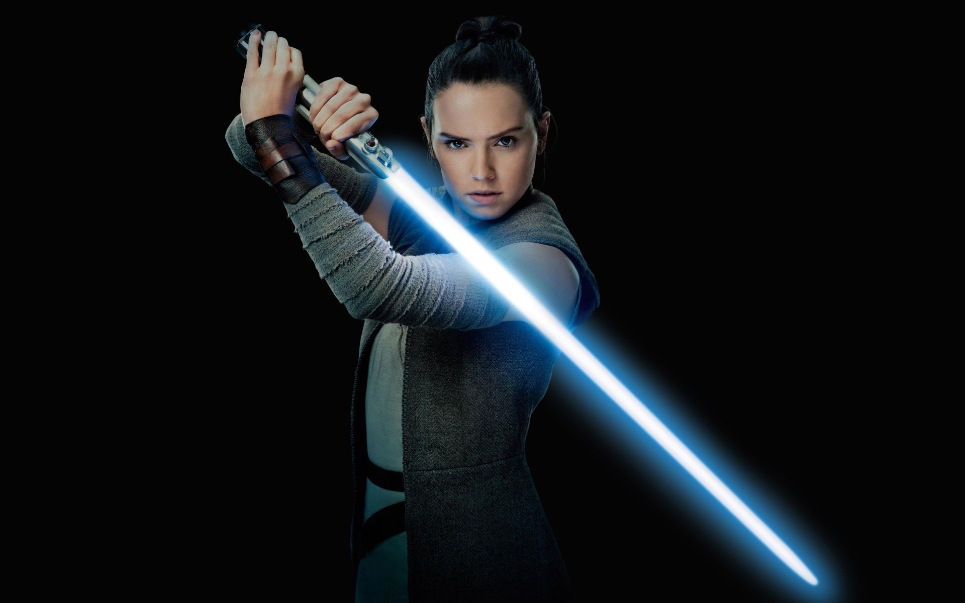 Rey from Star Wars: The Last Jedi holds a lit lightsaber in a focused stance, ready for battle. This HD desktop wallpaper captures Daisy Ridley as the determined Jedi against a dark background.