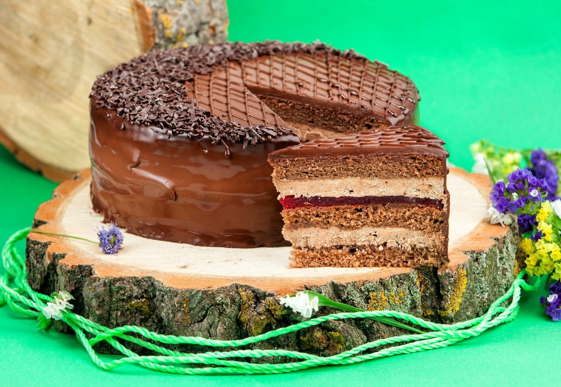 HD PC desktop wallpaper of a pastry/food cake: chocolate-layered cake with a sliced piece on a wooden slab, glossy glaze and shavings, purple flowers on a bright green background.