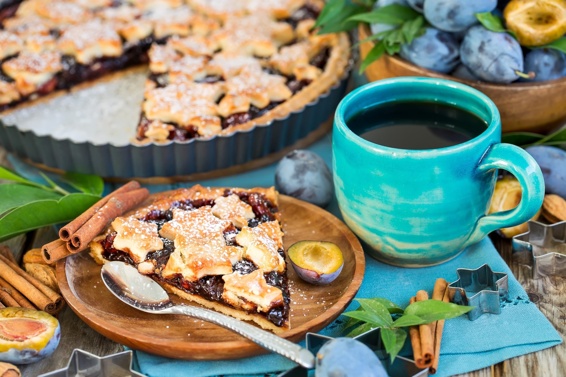 A 4K Ultra HD still life featuring a slice of plum pie dusted with powdered sugar, a blue cup of coffee, fresh plums, cinnamon sticks, and a pastry in the background.