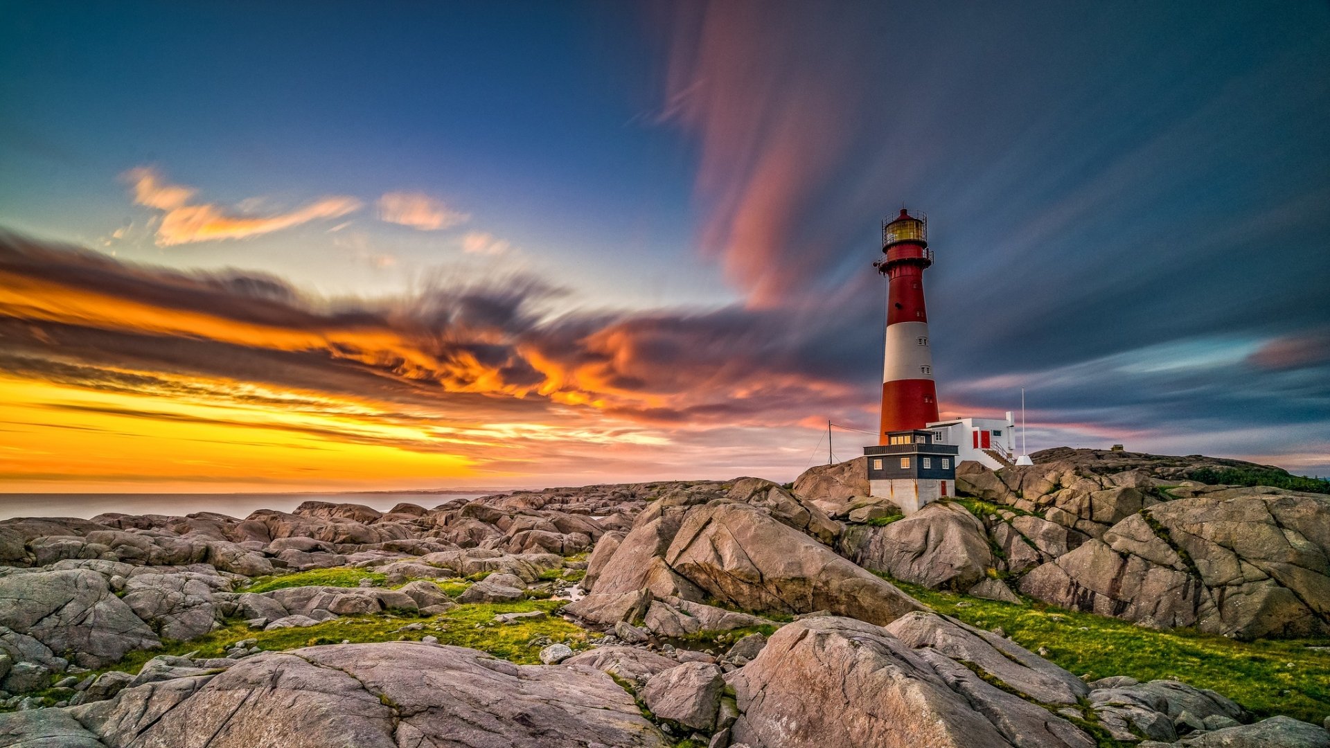 HD desktop wallpaper featuring a red and white lighthouse on rocky terrain by the ocean, beneath a vibrant sky with clouds at the horizon.