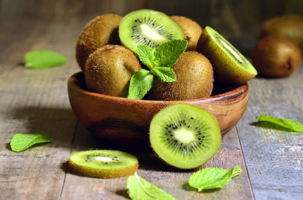A wooden bowl filled with whole and sliced kiwis on a rustic wooden surface, with fresh green mint leaves, captured in vivid 4K Ultra HD quality.