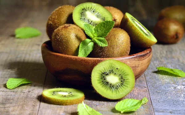 A wooden bowl filled with whole and sliced kiwis on a rustic wooden surface, with fresh green mint leaves, captured in vivid 4K Ultra HD quality.