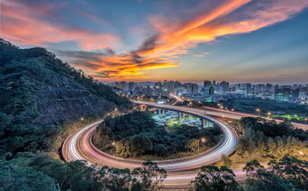 Time-lapse view of a bustling highway looping around greenery with skyscrapers under a vibrant sunset sky in a Taiwanese city, captured in HD.