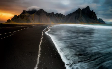 HD desktop wallpaper featuring Vestrahorn Mountain in Iceland. The image showcases dark sand, ocean foam, and a dramatic mountain backdrop at sunset.