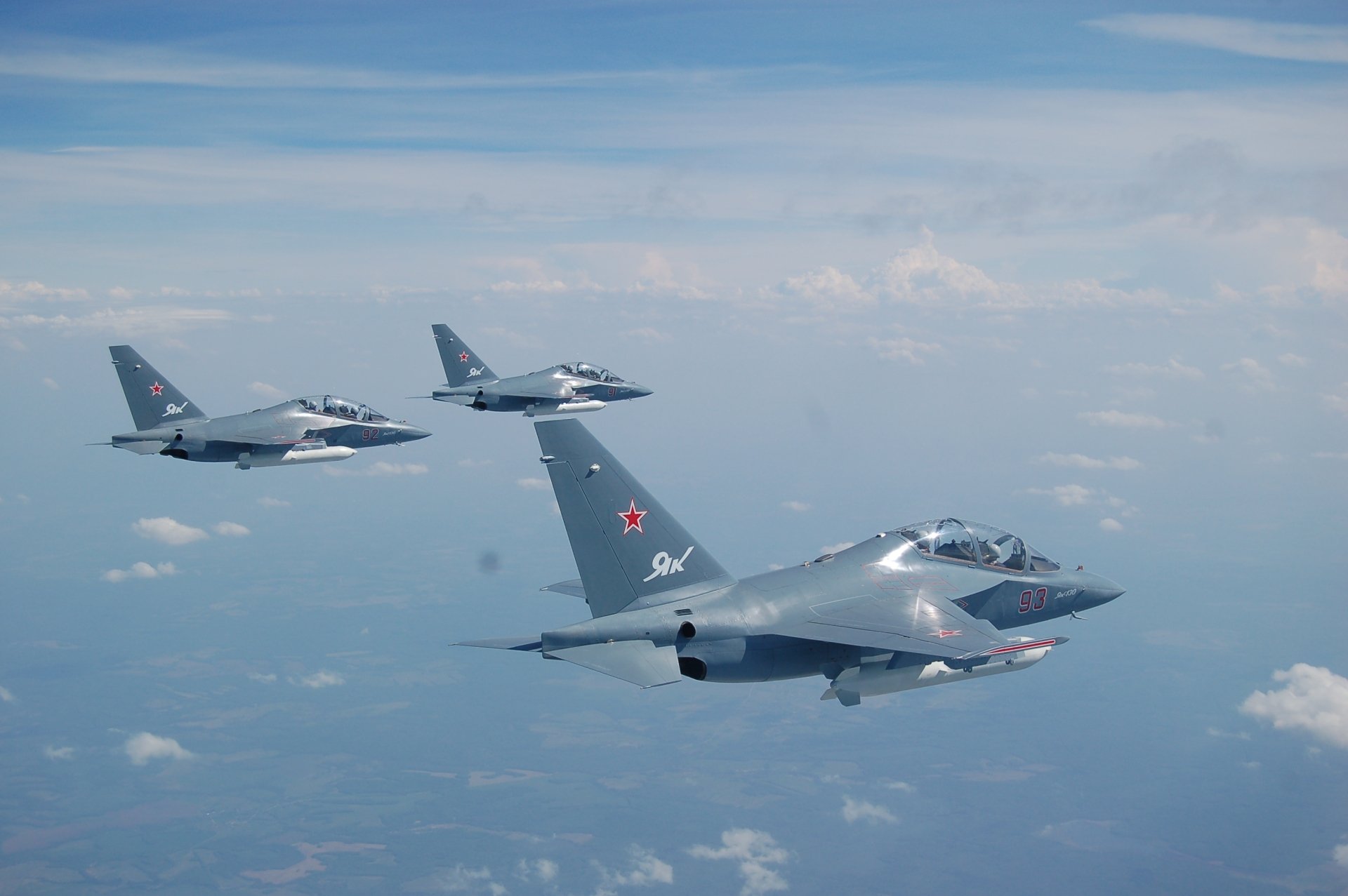 Three Yakovlev Yak-130 military jet fighter warplanes flying in formation above the clouds against a blue sky, captured in high-definition desktop wallpaper quality.