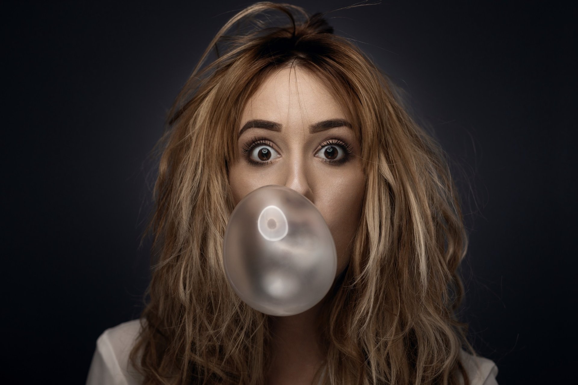 Close-up of a brunette woman with brown eyes blowing a bubble with chewing gum, set against a dark background for an HD desktop wallpaper.