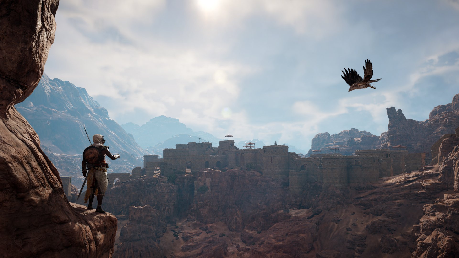 Bayek of Siwa stands on a cliff overlooking an ancient fortress under a bright sky, from the video game Assassin's Creed Origins in 4K Ultra HD.