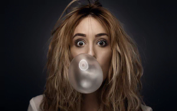 Close-up of a brunette woman with brown eyes blowing a bubble with chewing gum, set against a dark background for an HD desktop wallpaper.