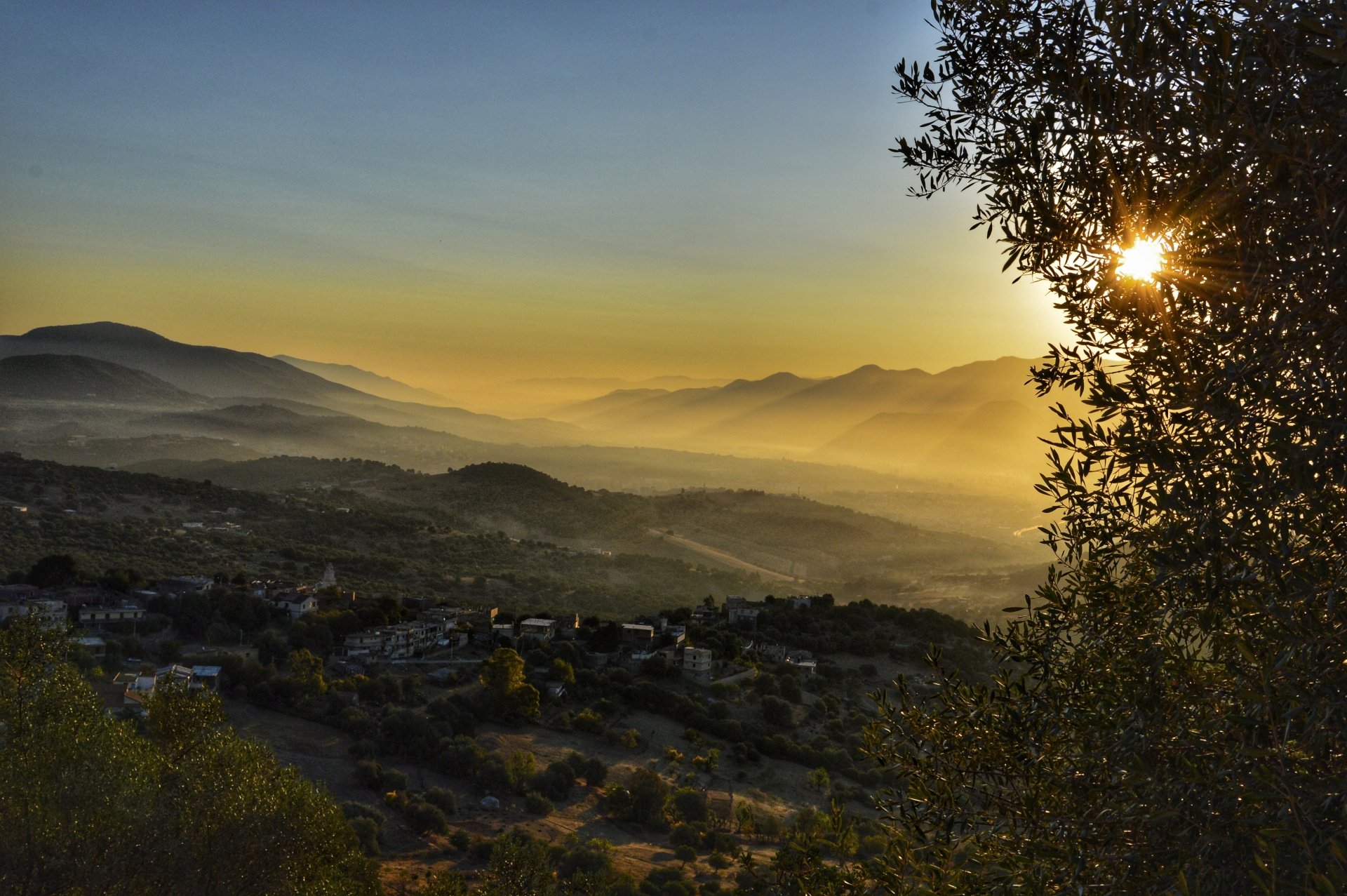 5K Ultra HD PC desktop wallpaper: sunrise over Bejaia mountains, misty valleys and a man-made village below, sun peeking through trees at right.