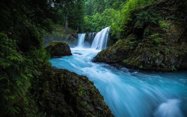 HD desktop wallpaper featuring a tranquil scene of a clear blue river cascading into a waterfall, surrounded by lush greenery and moss-covered rocks.