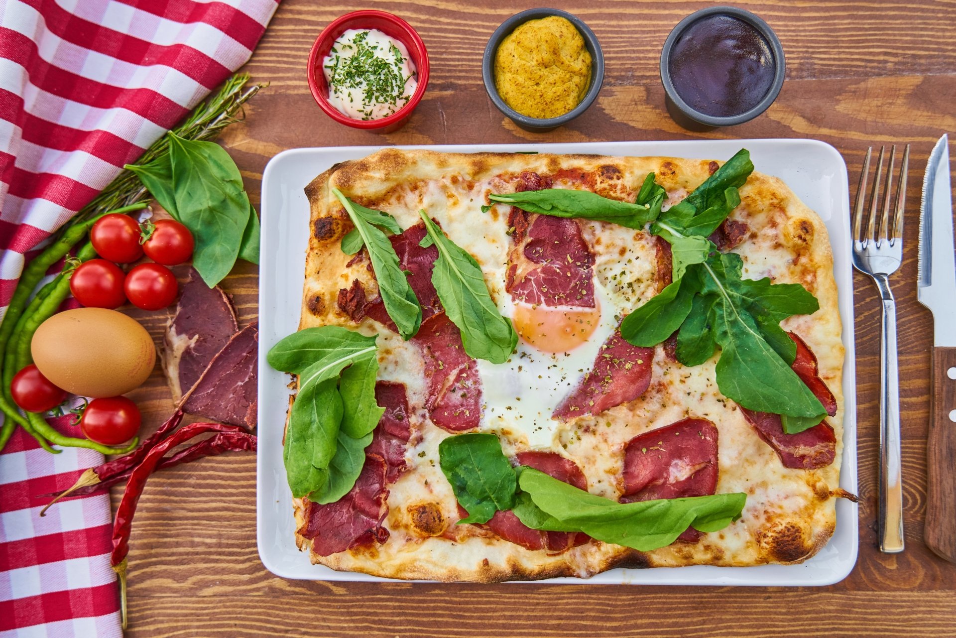 8K Ultra HD still life of a rectangular pizza topped with cured meat, fresh greens, and an egg, surrounded by sauces and fresh ingredients on a wooden table.