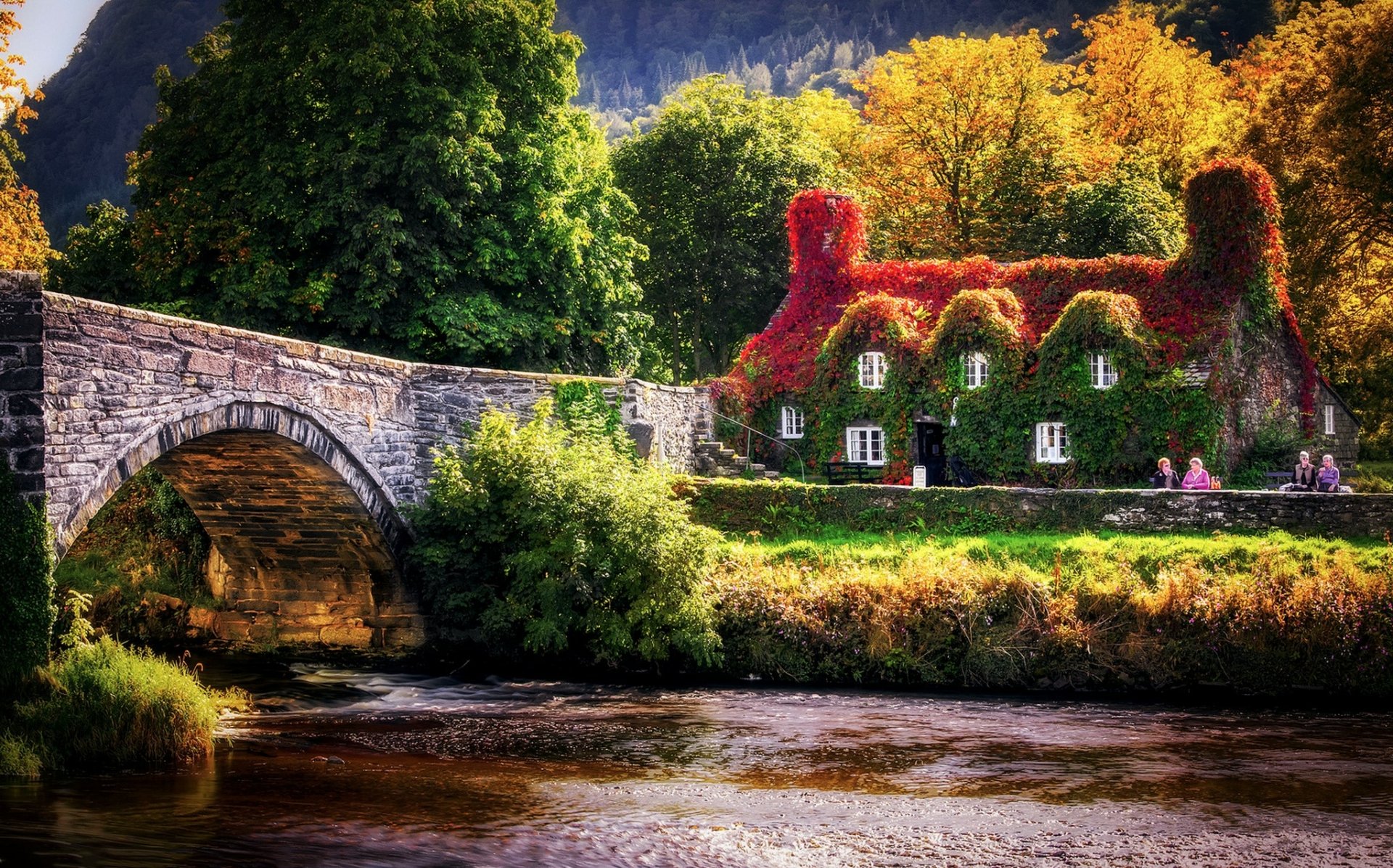 Autumn scene in Wales featuring a stone bridge over a river, with a house covered in red ivy nestled among colorful fall foliage.