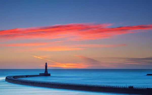 HD desktop wallpaper featuring a serene ocean view at sunset, with a vibrant sky and a lone lighthouse on a curved pier.