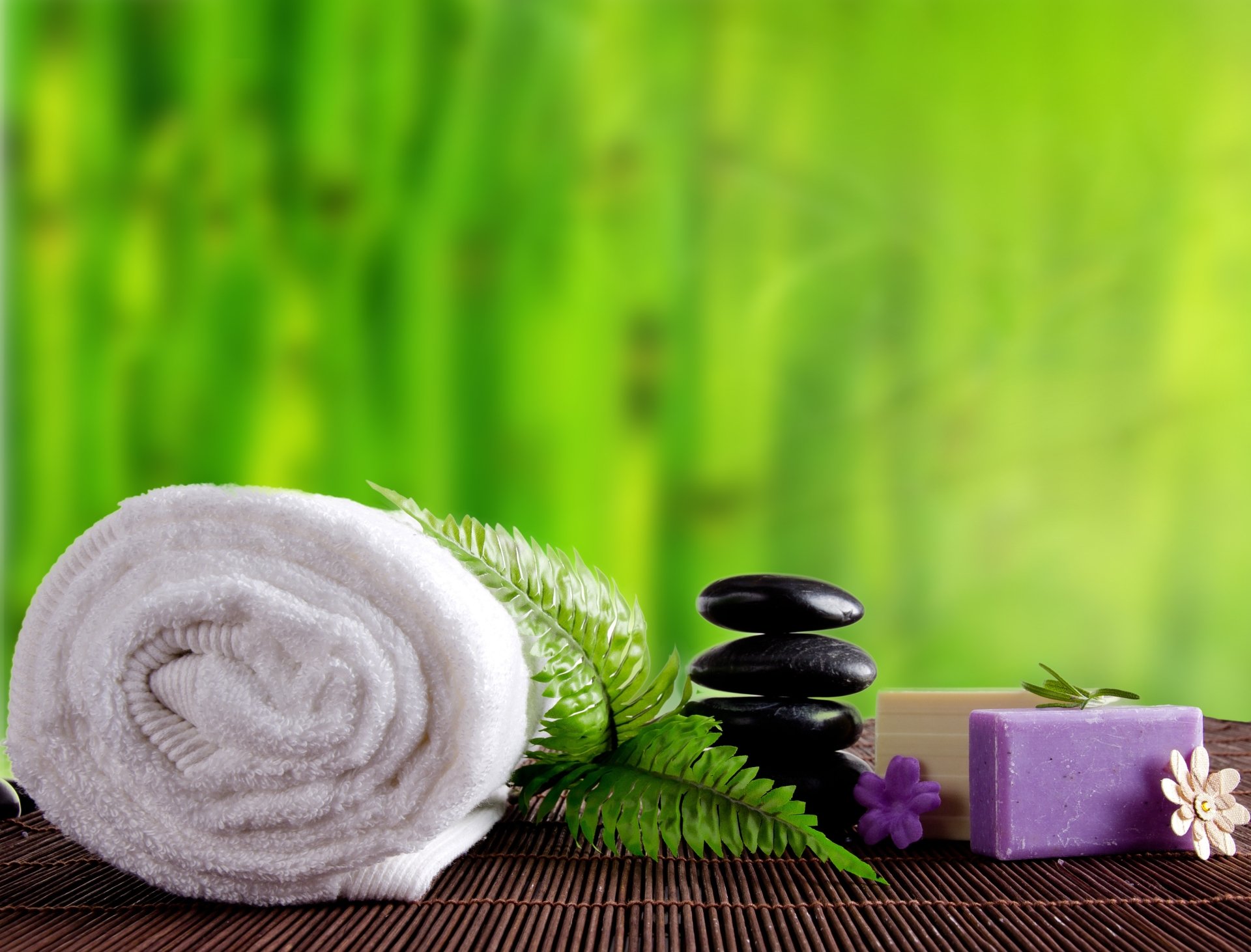 A serene spa scene with stacked black stones, a rolled white towel, soap bars, and green fern leaves against a vibrant bamboo background in 4K Ultra HD.
