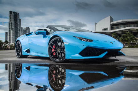 HD desktop wallpaper featuring a blue Lamborghini Huracan supercar, with its reflection visible on wet pavement, set against a backdrop of modern city buildings.