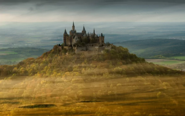 HD wallpaper featuring Hohenzollern Castle in Germany, perched atop a hill with lush, golden foliage surrounding the majestic, historic structure against a backdrop of expansive landscape and moody sky.