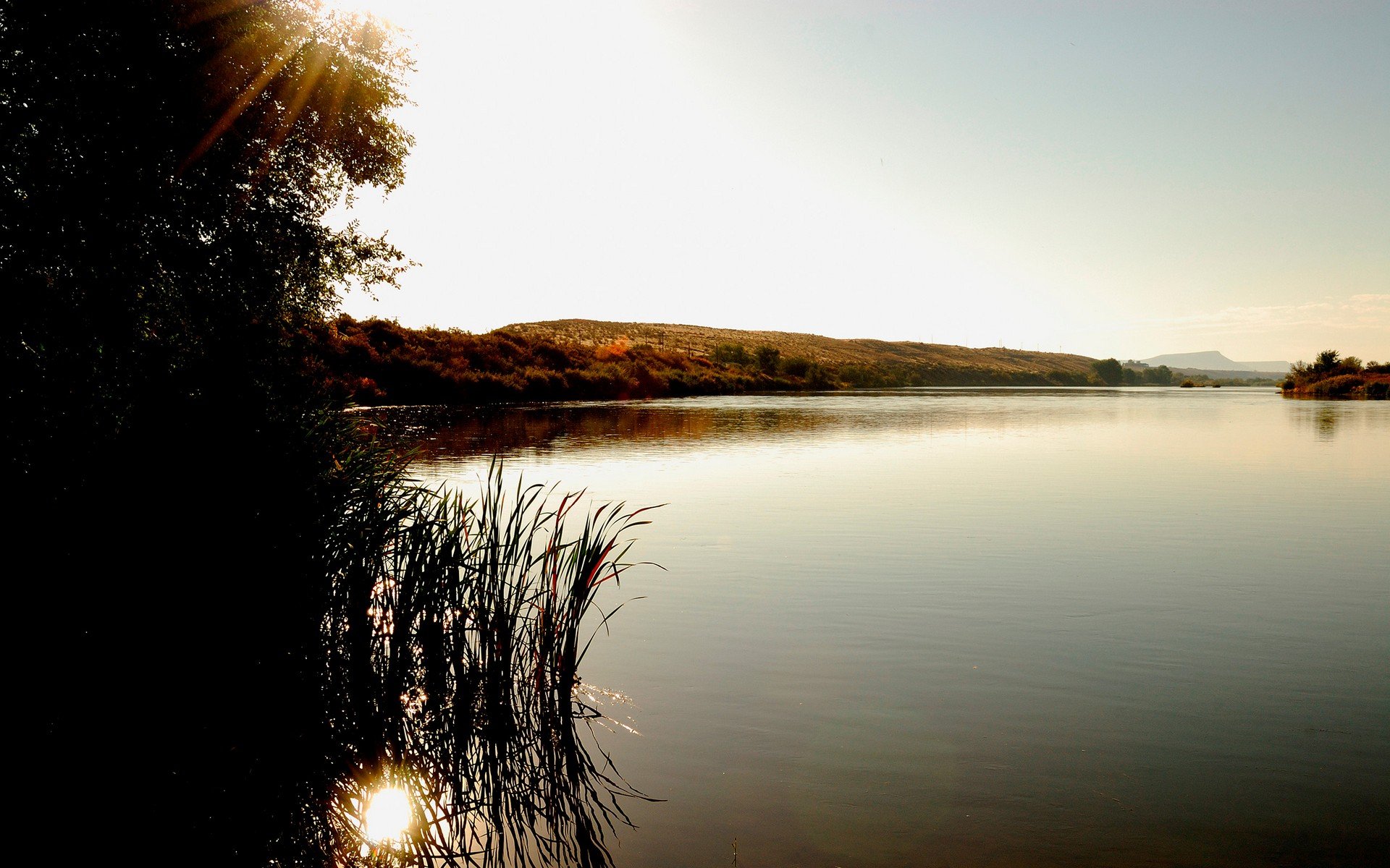 HD desktop wallpaper of a serene lake at sunrise, with sunlight reflecting on calm water surrounded by grass, bushes, hills, and an expansive sky in a peaceful wilderness landscape.