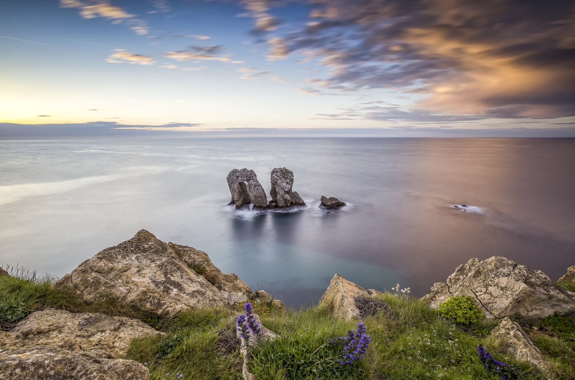 A natural rock arch rises from the ocean under a vibrant sky at the horizon, captured in a high-definition desktop wallpaper showcasing stunning nature scenery.