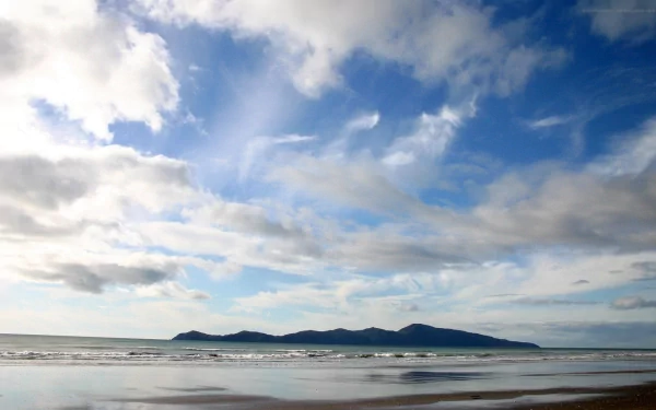 A serene beach landscape features gentle waves lapping at the shore, with a sunny sky and clouds above. In the distance, a mountain island rises above the sparkling sea.
