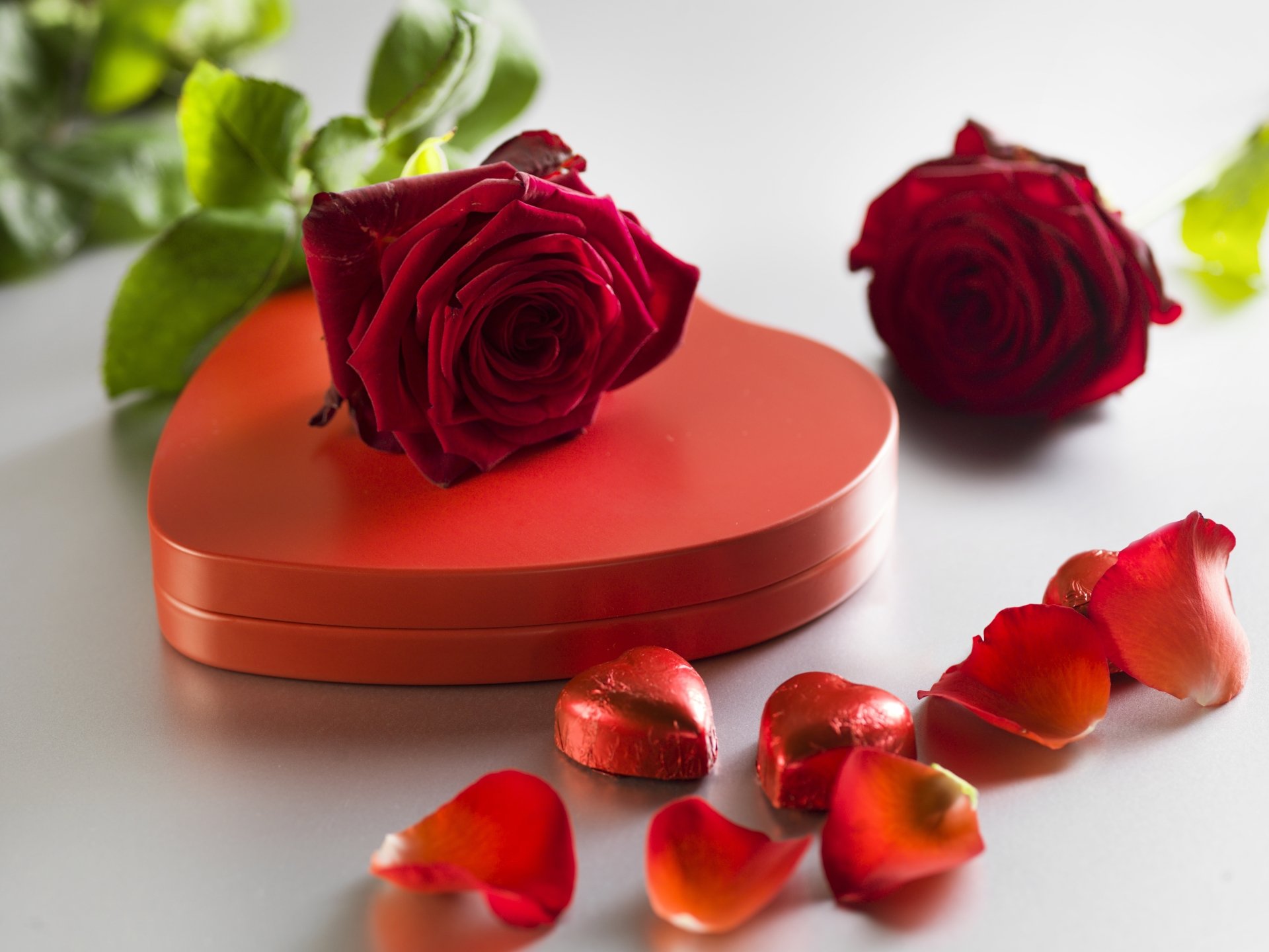 A 4K Ultra HD desktop wallpaper featuring two red roses, heart-shaped chocolates, and scattered rose petals, evoking a romantic Valentine's Day holiday theme.