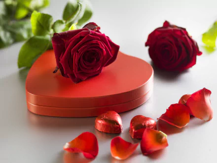 A 4K Ultra HD desktop wallpaper featuring two red roses, heart-shaped chocolates, and scattered rose petals, evoking a romantic Valentine's Day holiday theme.