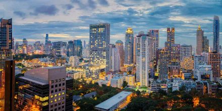 Nighttime cityscape of Bangkok, Thailand showcasing illuminated skyscrapers and buildings under a cloudy sky, captured in high definition.