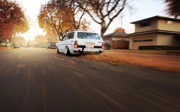 White Datsun 610 Wagon driving down a suburban street with autumn trees, captured in a dynamic motion shot for an HD PC desktop wallpaper background.