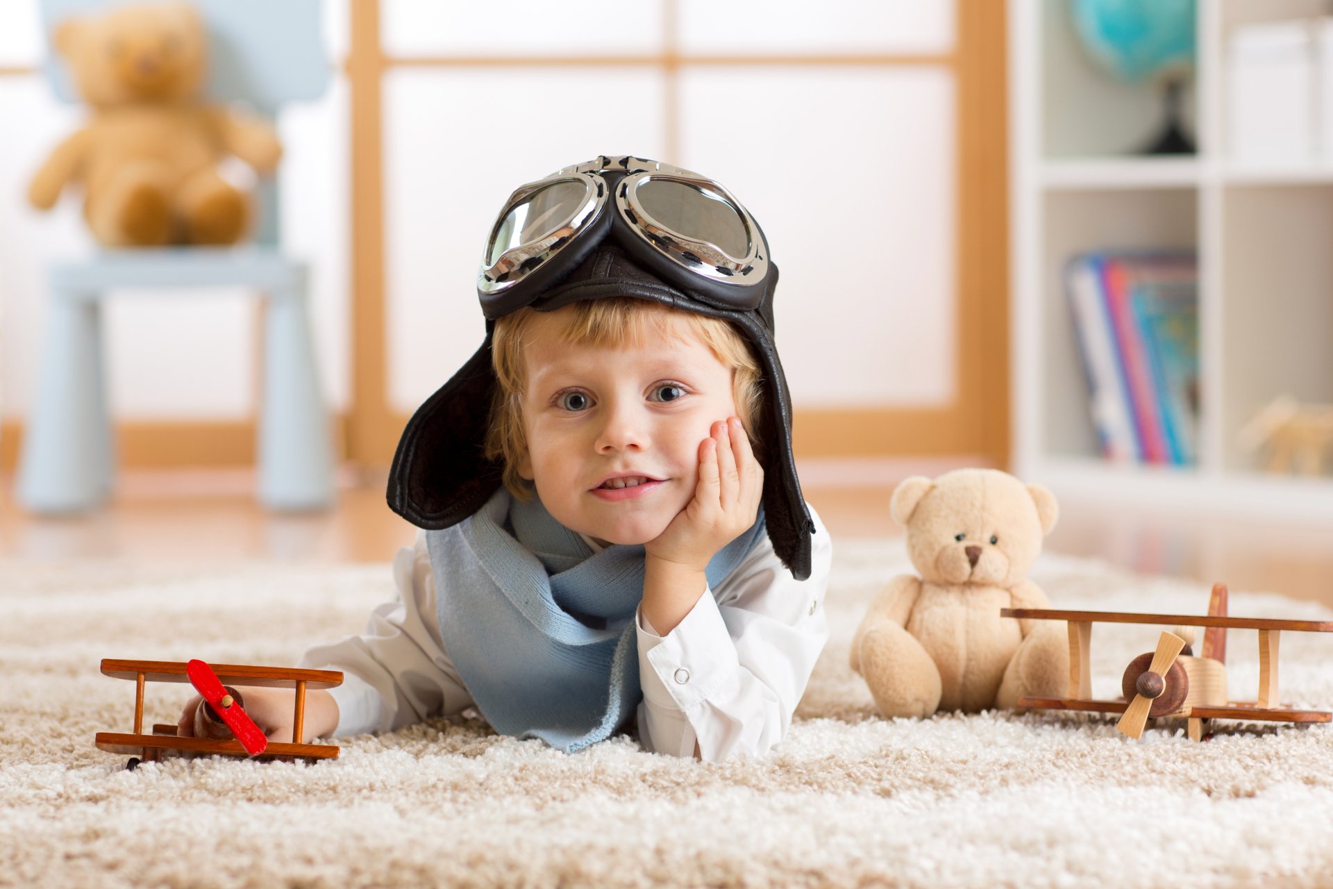 A little boy with blue eyes wearing an aviator helmet lies on the floor surrounded by toy planes and a teddy bear, captured in sharp 4K Ultra HD with depth of field.