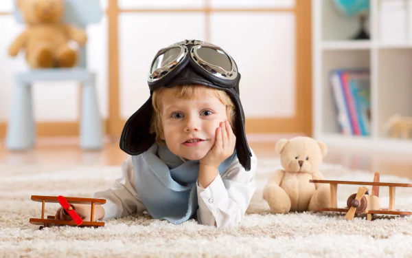 A little boy with blue eyes wearing an aviator helmet lies on the floor surrounded by toy planes and a teddy bear, captured in sharp 4K Ultra HD with depth of field.
