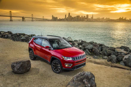 Red Jeep Compass SUV parked on a rocky waterfront with city skyline and bridge at sunset — 2K Quad HD PC desktop wallpaper background.