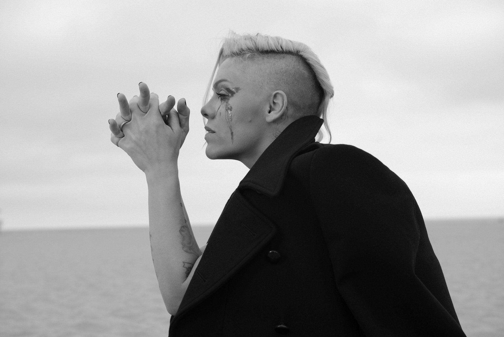 Black-and-white monochrome 4K Ultra HD desktop wallpaper: profile portrait of an American singer with shaved-side haircut and dramatic eye makeup, hands clasped, set against a calm seaside backdrop.